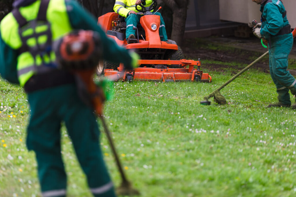 Three landscapers in uniform mowing and cleaning a green grass area with lawn equipment